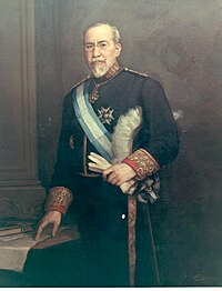 Standing portrait of 60-ish man with a white beard, mustache, and receding hairline, in circa-1900 formal dress with glasses, a high collar, a star-shaped badge on his jacket, and a blue and white sash, holding a glove and loosely pointing to a paper on a table.