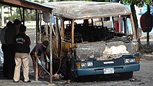 A color photograph of various individuals inspecting a charred minibus which was the target of an arson attack