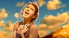 A brunette teen tilts her head upward and looks to the sky while clasping her hands at her chest and singing: She sits at the top of a red mountain cliff during sunset.