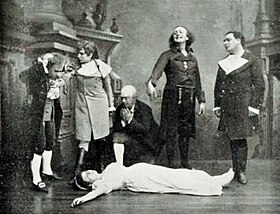 Photograph of white man with long hair in 19th century day clothes gesticulating manically over a female corpse, while two other men and a woman look on, horror-struck