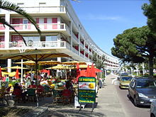 Photographie présentant un long bâtiment d'habitations de forme courbe et abritant en terrasse des restaurants et commerces touristiques au bord d'une rue très fréquentée