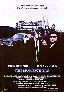 Movie poster with two of the main characters on the right-side of the image. They are both wearing black suits, hats, and sunglasses and facing forwards. The man on the right is resting his arm on the shoulder of the man on the left. A police car is present on the left side of the image behind them. At the top of the image is the tagline "They'll never get caught. They're on a mission from God." At the bottom of the poster is the title of the film, cast names, and production credits.