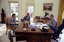 A man sits at his desk, smoking a pipe, while two other men speak to him from the other side of the desk.