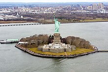 A tall green statue on an island in a harbor.