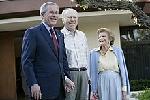 President Bush in a suit standing next to the Fords in casual attire in front of their yellow house.