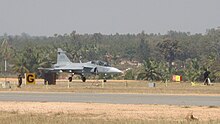 Jet aircraft in the distance preparing to take off from rural airport surrounded by green trees