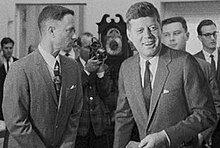 Black-and-white film screenshot showing the main character on the left looking toward another man, President Kennedy, (voiced by actor Jed Gillin), on the right. Kennedy is smiling and looking to his left. In the background, several men are looking in different directions and one is aiming a camera.