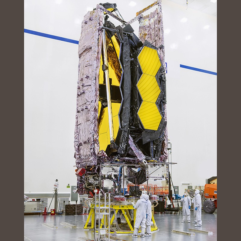 An instrument with gold hexagonal mirrors, wrapped in shiny material and attended by people in clean suits.