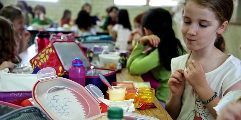 The picture shows a group of children in a school canteen eating food from their lunch boxes.