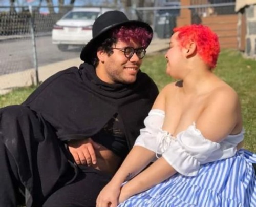 a photo of Beryl, a lightskinned, fat Afrolatine trans woman, and Gardenia, a lightskinned, mixed Black tme nonbinary person. Beryl's curly hair is short on the sides and long on top, and the top is dyed purple. she is wearing a black wide brimmed hat, a black cape with a hood, a black crop top with a hand-painted gold design on it, and black sweatpants. Gardenia's hair is cut very short, and is dyed hot pink with magenta cow spots. he is wearing a white sleeved tube top  and a blue and white striped skirt. Beryl is lounging in a reclined position, and Gardenia is sitting with his legs underneath him. both are smiling, and looking at eachother with a loving expression. in the background, a yard fence is visible, as is the side of an apartment building, a white car, and another next door building.