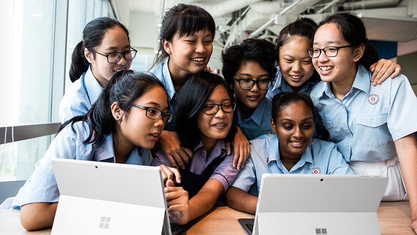 A group of girls sit and stand in front of two Microsoft tablets.