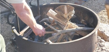 A hand reaches into a metal campfire ring to rearrange the wood