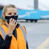 Ramona Moraru, supervisora de operaciones en la estación logística de Getafe. En el exterior del centro logístico con dos camiones de Amazon Prime de color azul con la sonrisa de Amazon en blanco. Ramona está hablando por un walkie talkie. Tiene el pelo claro, recogido en una cola, Con la mano derecha agarra el walkie talkie, tiene las uñas pintadas de negro. Lleva un tatuaje en la muñeca izquierda y un pulsera. Los ojos oscuros y maquillados y una mascarilla negra de Amazon. Lleva un chaleco de color naranja y un jersey negro con estrellas de color gris.  