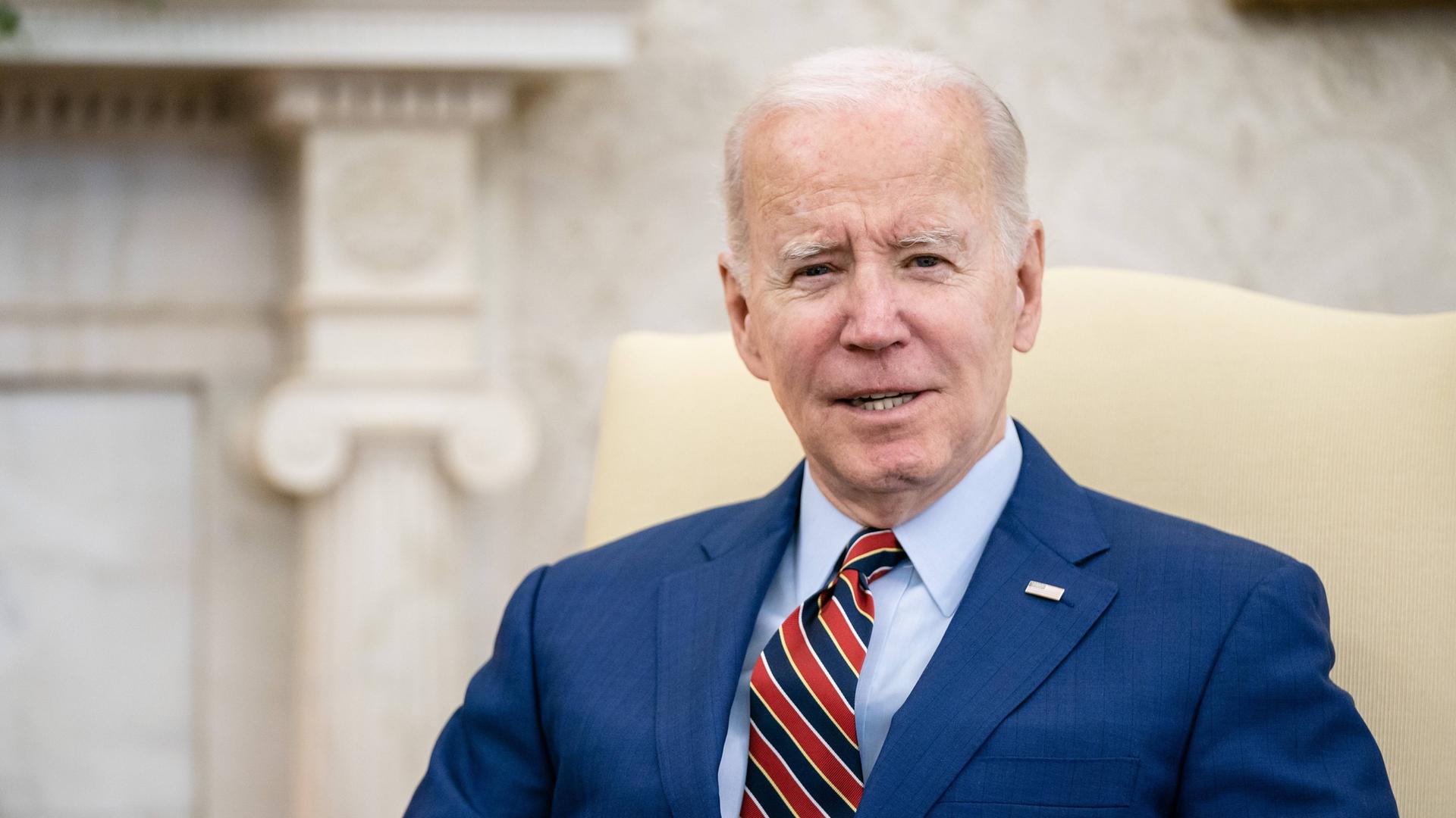 Joe Biden sitzt auf einem weißen Sessel, trägt ein blaues Sacko und schaut freundlich in die Kamera