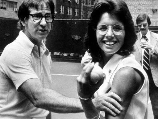 Tennisspieler Bobby Riggs fasst Tennisspielerin Billy Jean King spielerisch an die Armmuskeln, während Billy Jean King mit einem triumphierenden Lachen einen Tennisball in die Kamera hält.