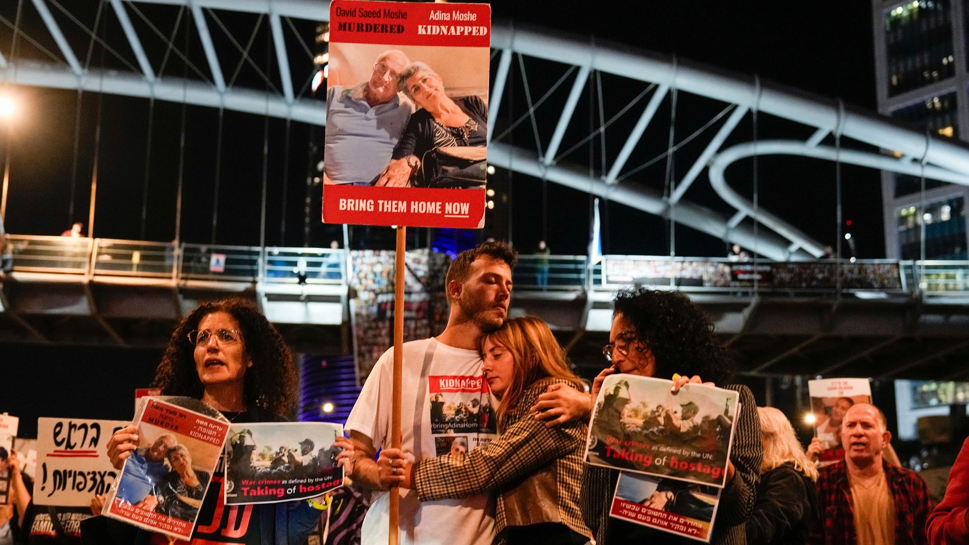 Menschen halten Plakate ihrer Angehörigen hoch. Ein Paar steht in der Mitte des Bildes und umarmt sich.