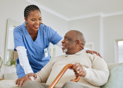 An elderly Black man wearing a tan sweater and seated holding a cane while looking up at a Black woman nurse with cornrows and wearing blue scrubs while resting her hand on the man's shoulder.