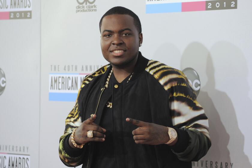 Sean Kingston in a black shirt in a tiger-print jacket pointing up his two finger posing against a white backdrop