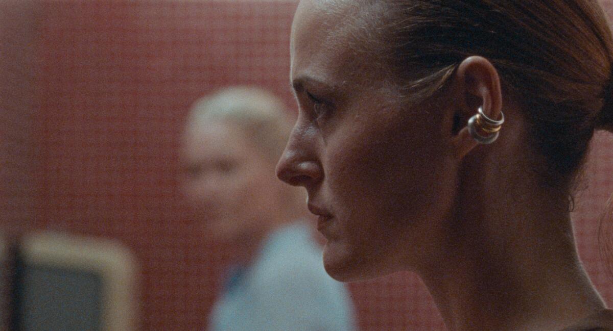 A close-up of a woman wearing earrings in profile, as another woman out of focus glares at her.