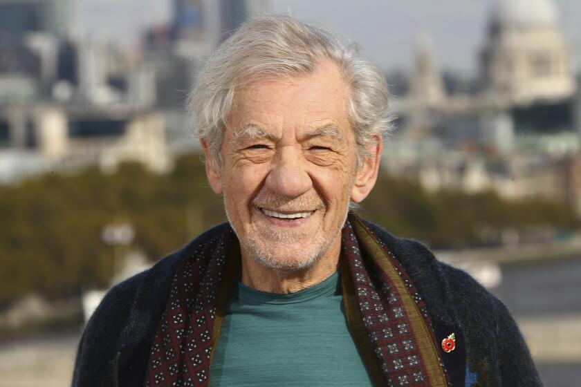  Ian McKellen wears a blue t-shirt and a printed scarf with a cardigan while posing in outdoors in front of a cityscape