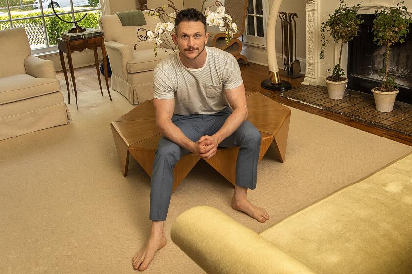 Actor Jonathan Tucker sits on a wooden coffee table near a fireplace, sofa and chairs in a beige-toned living room