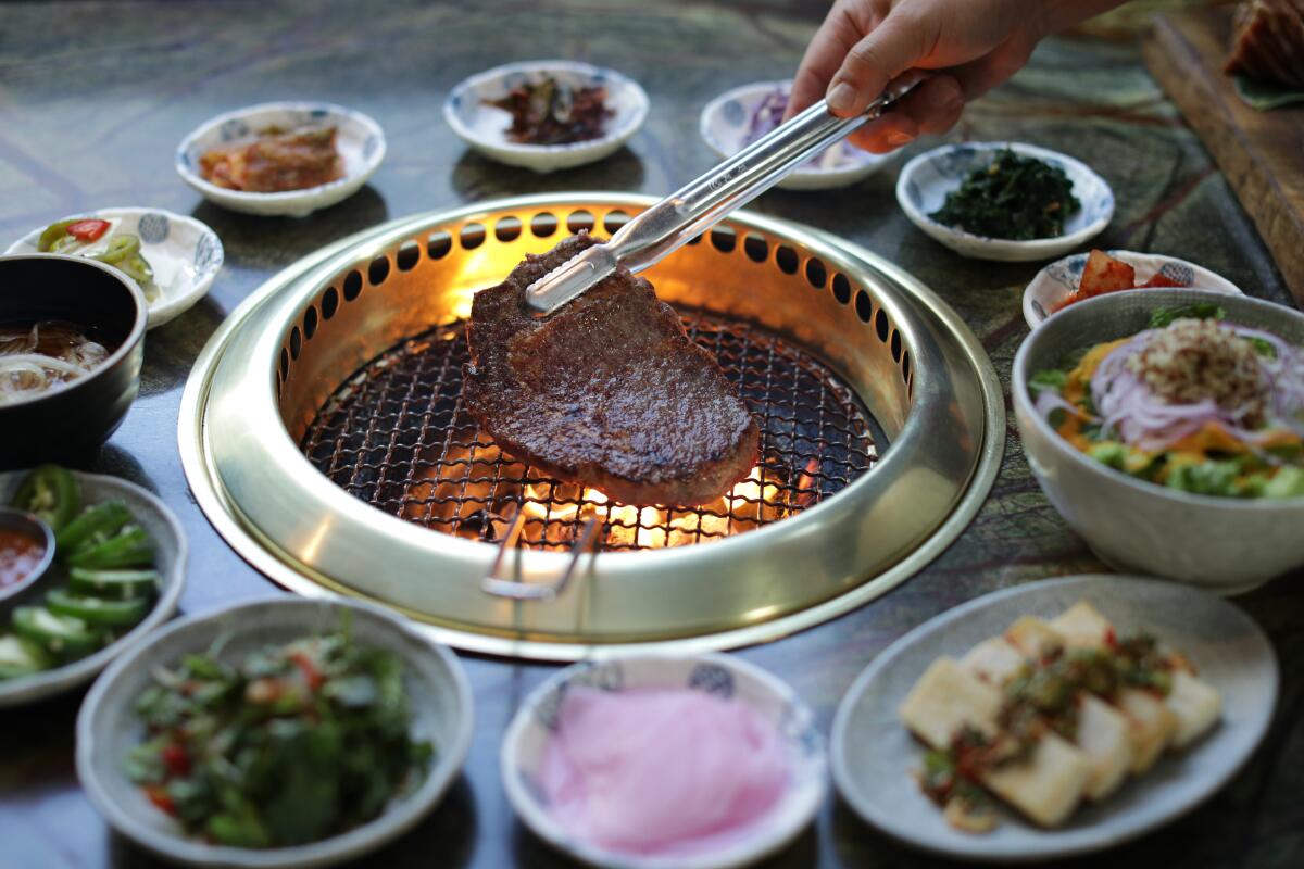 Tongs hold a rib-eye on the round metal grill at Jeong Yuk Jeom, surrounded by small side dishes.