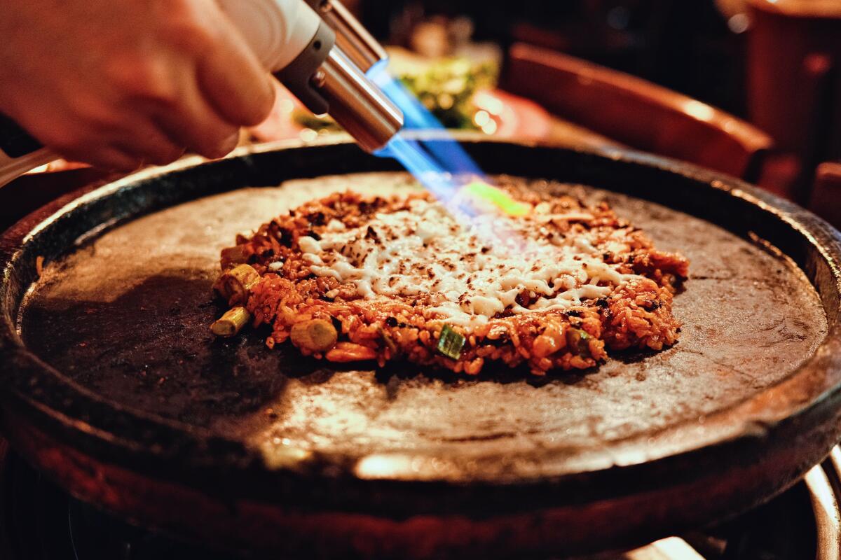 A Korean barbecue grill featuring a circle of kimchi fried rice. Two hands aim blowtorches at mozzarella in the center.