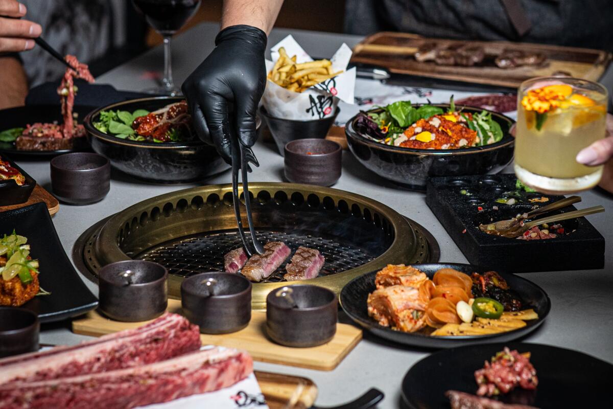 A black-gloved hand arranges meat on a grill, surrounded by various side dishes and sauces