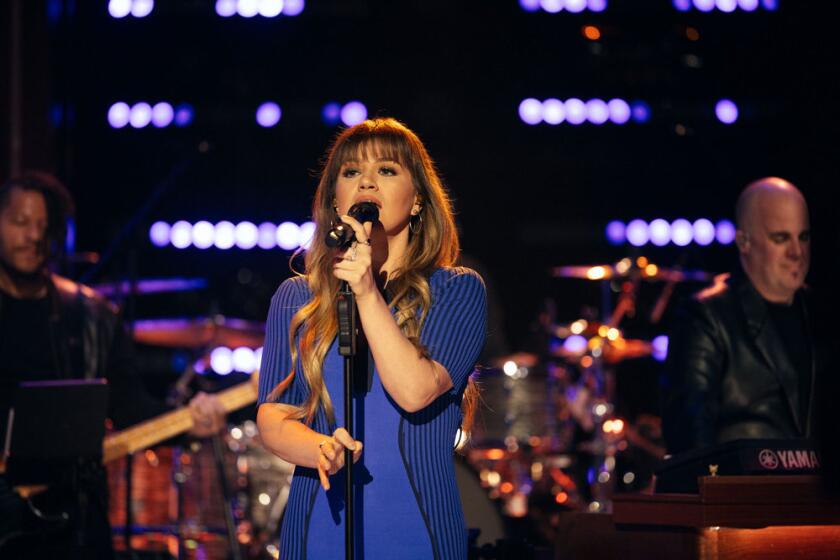 Kelly Clarkson in a blue dress with long wavy hair and bangs singing into a microphone 