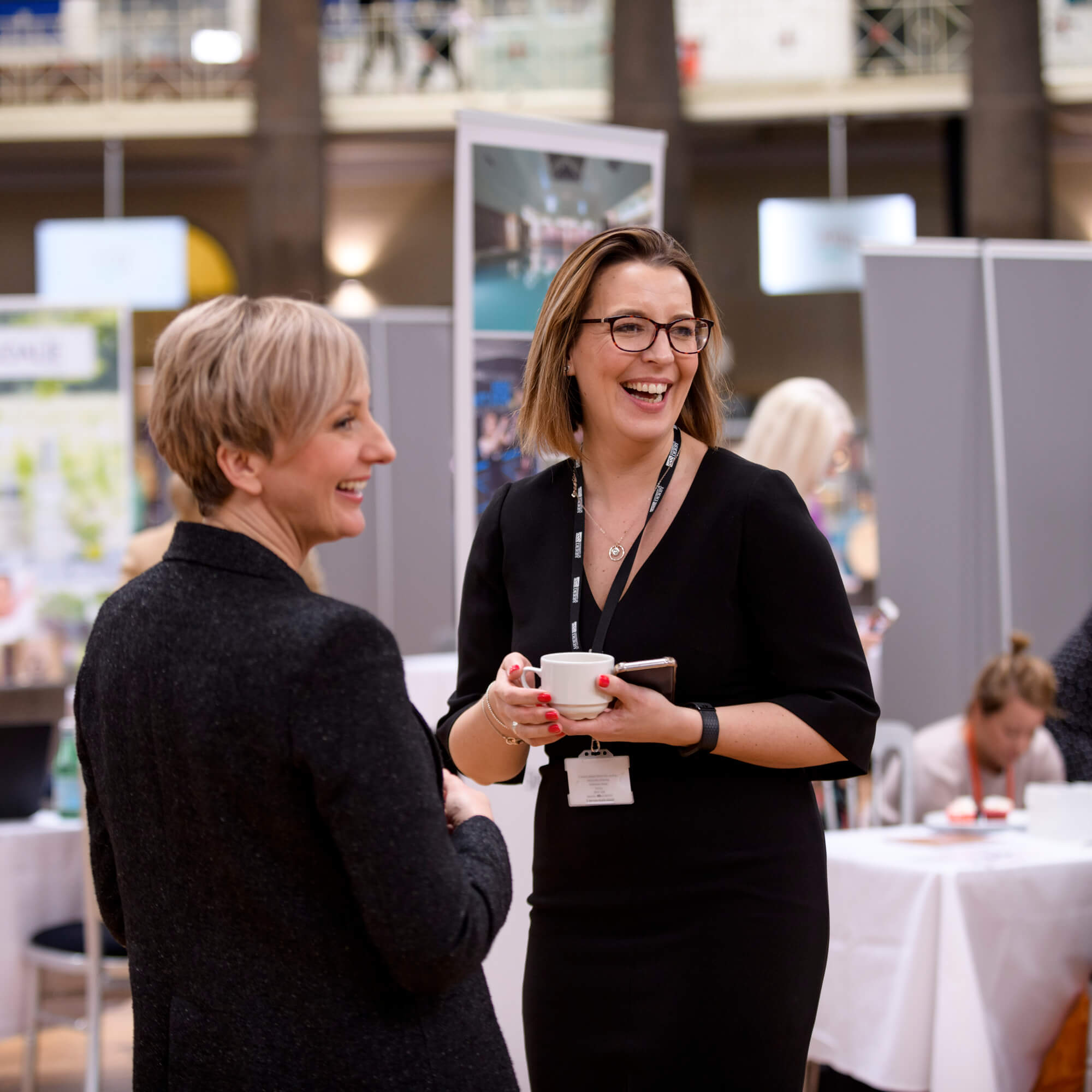 A woman holding a cup of tea smiling and talking to another woman who is also smiling in a professional networking setting