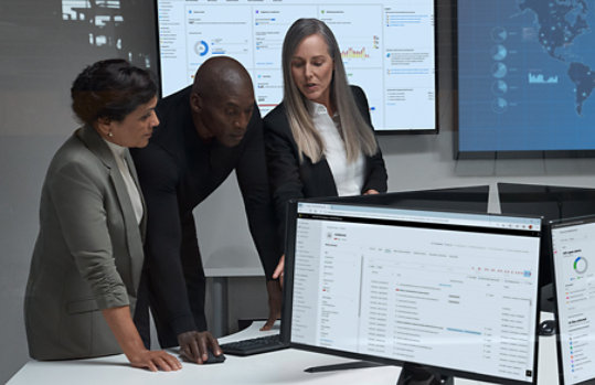 Three technology workers looking at a computer screen in an office environment with surrounding monitor screens displaying reports and maps.