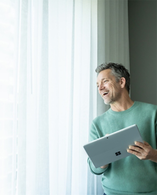 A man standing, smiling, and looking out a window while holding a Surface device