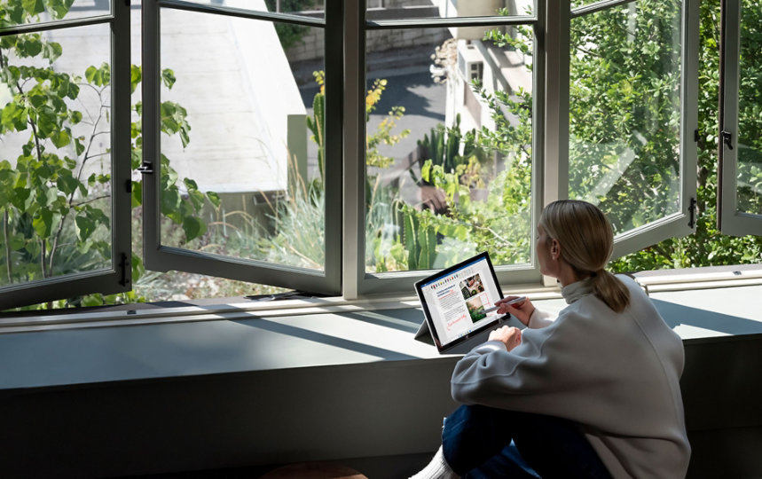 A woman sitting near bright open windows with a view of green plants and working on a Microsoft Surface Laptop.