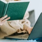 A student reads a book while a laptop rests on her legs as she studies for the SAT and ACT