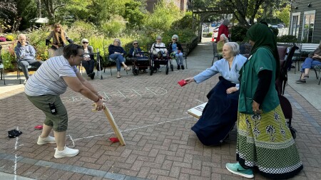 A woman in a wheelchair throws a cornhole bag towards a wooden platform being held up by a woman. A staff member in a green uniform and colorful skit stands next to her. A group of people watch, sitting in chairs.