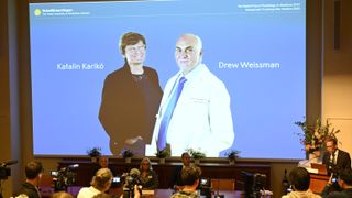 Thomas Perlmann, secretary of the Nobel committee, stands next to a large projector screen displaying the names and photos of Katalin Karikó and Drew Weissman