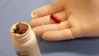 Gloved hand holding the antiviral molnupiravir, a red pill, next to a bottle of the drug