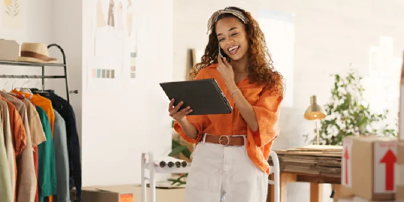 A woman in an orange shirt smiling while holding a tablet with a phone to her ear, surrounded by apparel for commerce platforms.