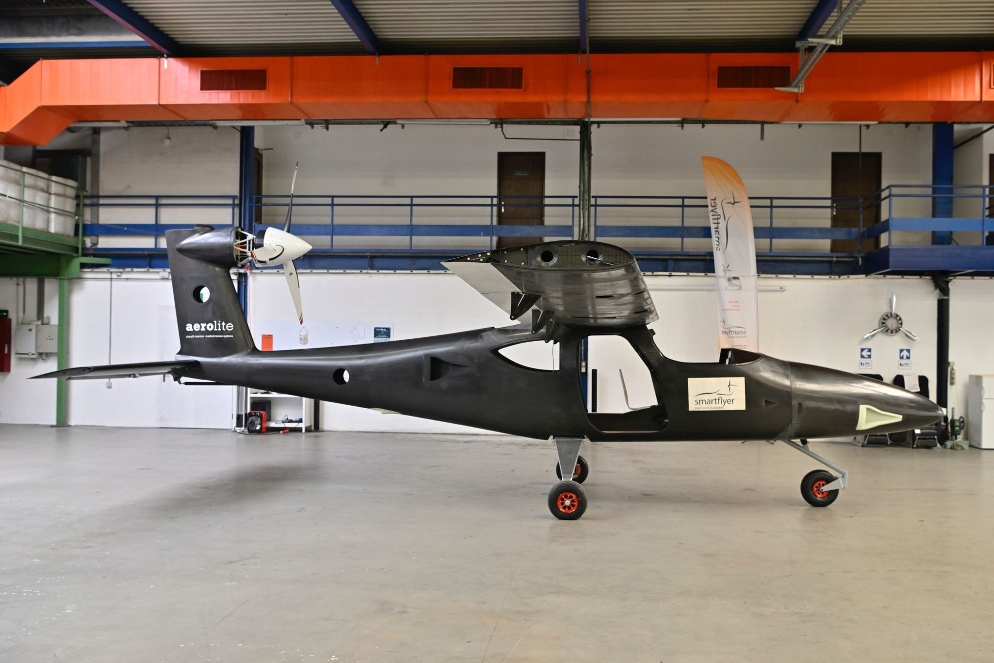 Prototyp eines autonom fliegenden Flugzeugs in einer Industriehalle aufgebockt, mit Propeller am Heck und auffälligem Design in Schwarz und Orange.