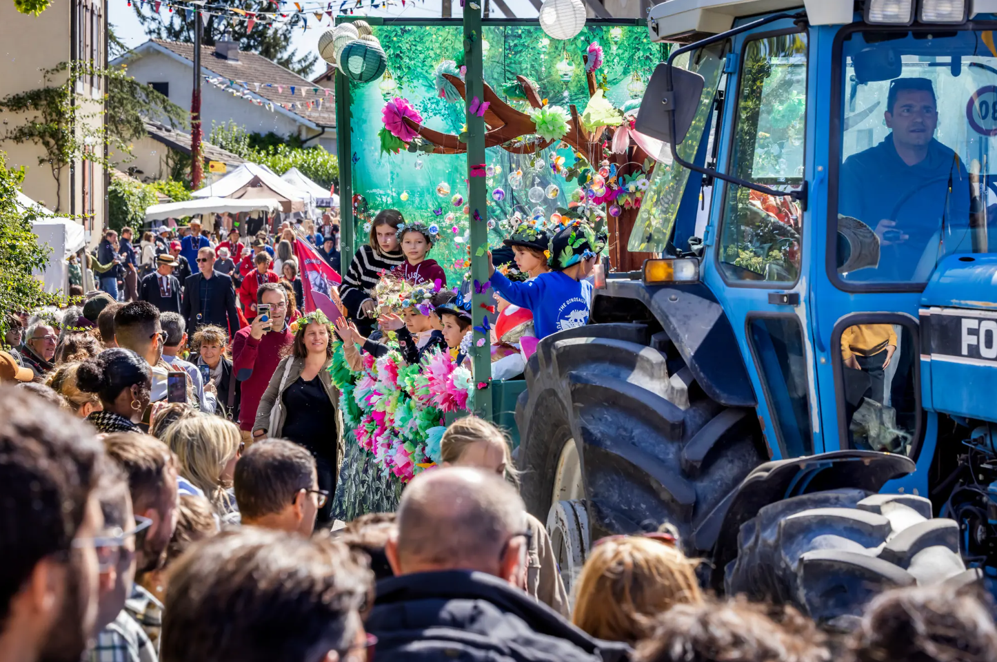 Un tracteur bleu décore avec des fleurs colorées lors d'un défilé de fête, entouré de nombreux spectateurs dans une rue.