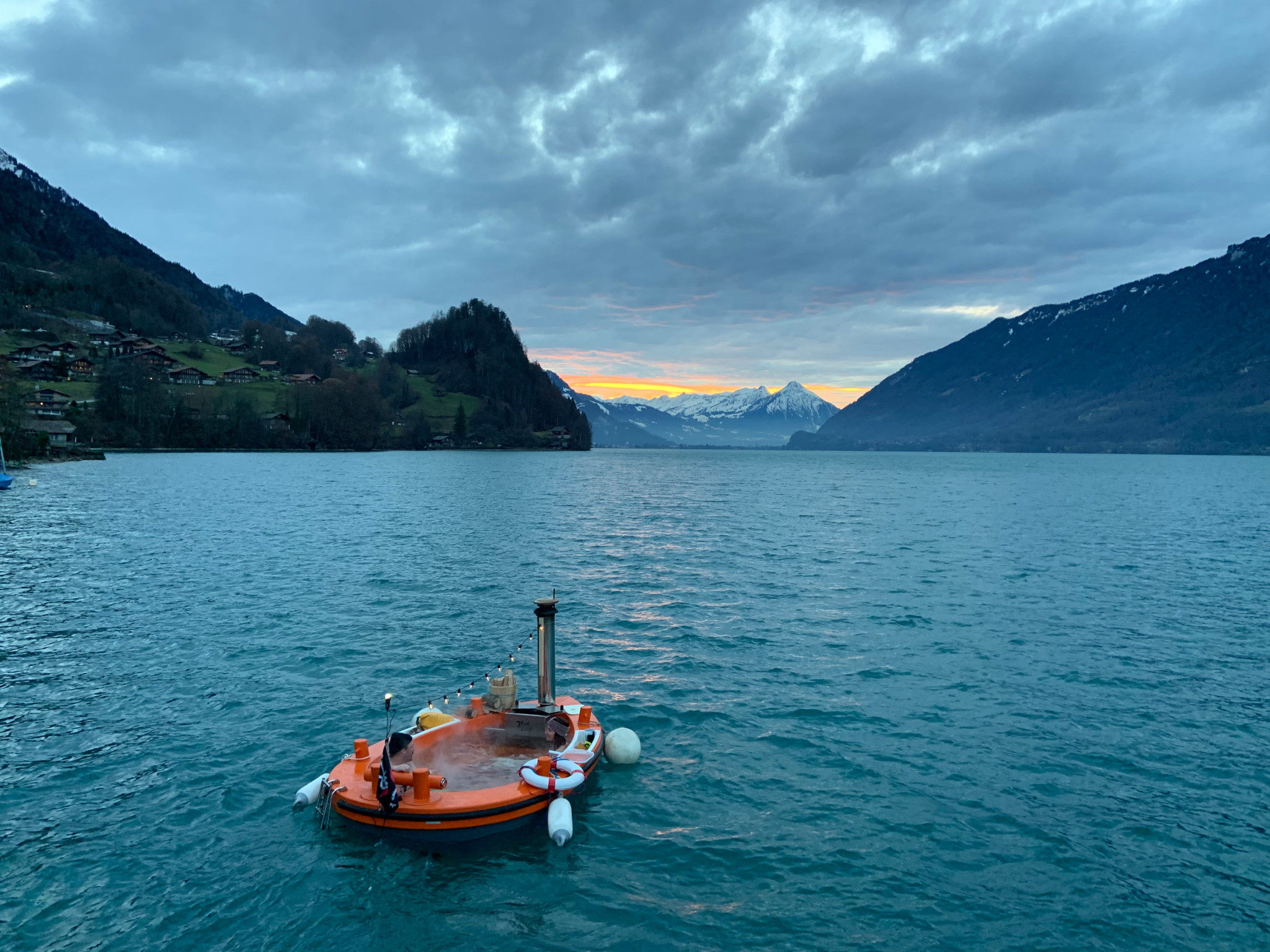 Ein Hot Tug, ein schwimmender Whirlpool, fährt auf dem Brienzersee bei Sonnenuntergang mit schneebedeckten Bergen im Hintergrund.