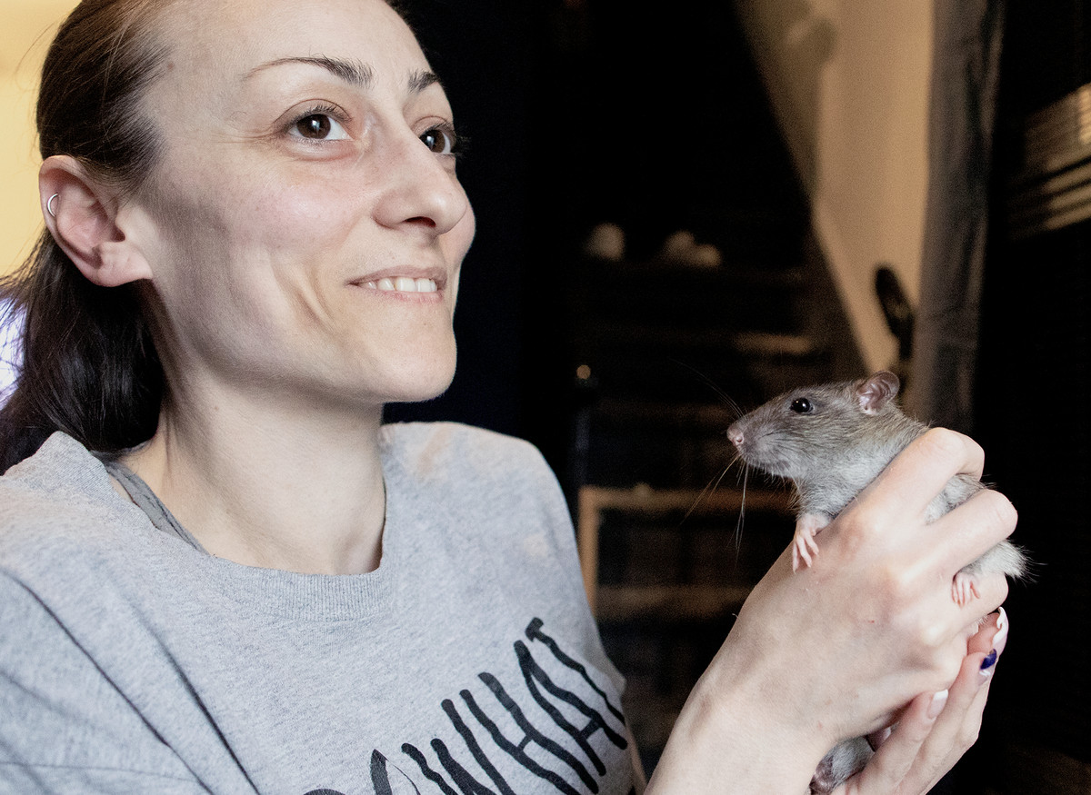 A woman with dark hair and a gray T-shirt smiles while holding a rat with two hands in front of her.