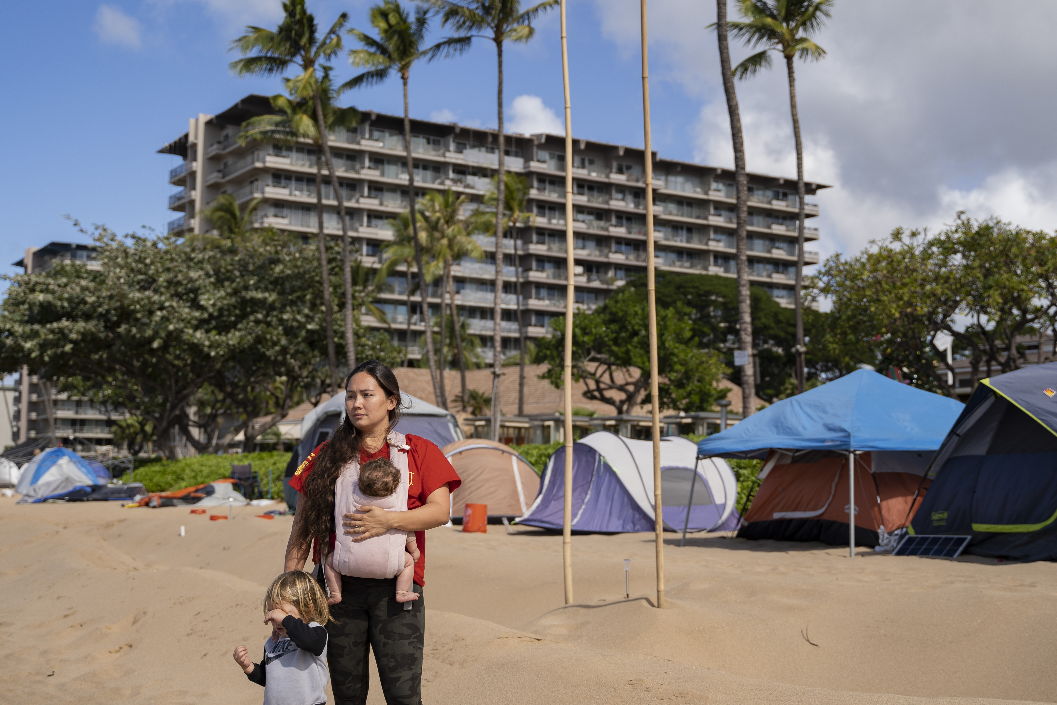 A woman with two young children stands on a beach in front of a multistory hotel, palm trees, and tents.