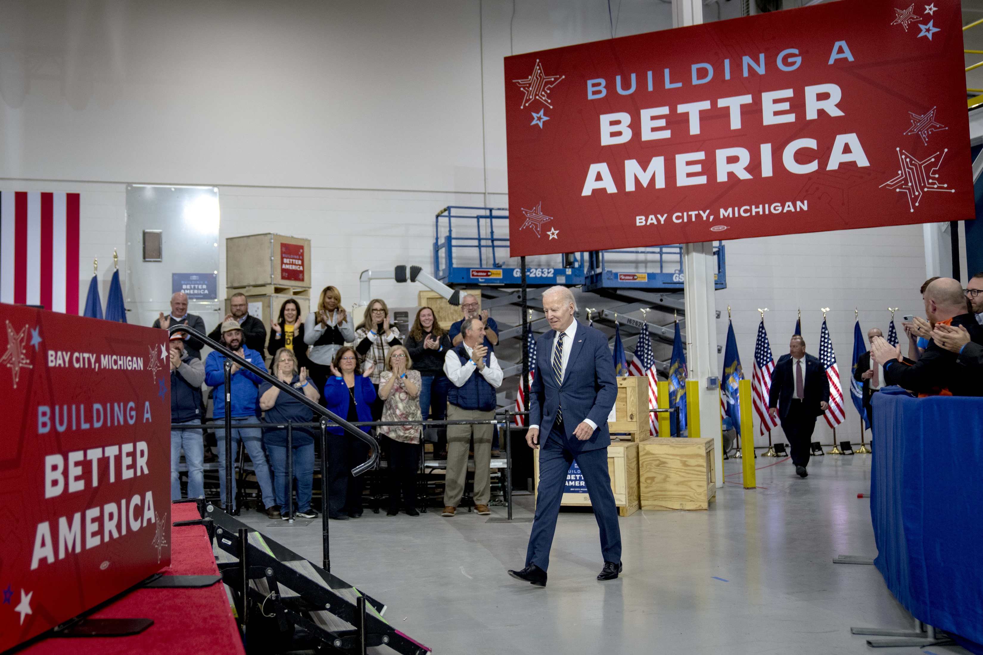 Joe Biden, pictured mid-stride, walks under a large red banner that reads: “Building a Better America.” A small group of people look on and clap.