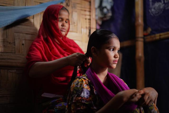 2 girls sitting down. 1 girl with a red headscarf is modeling the other's girl hair.