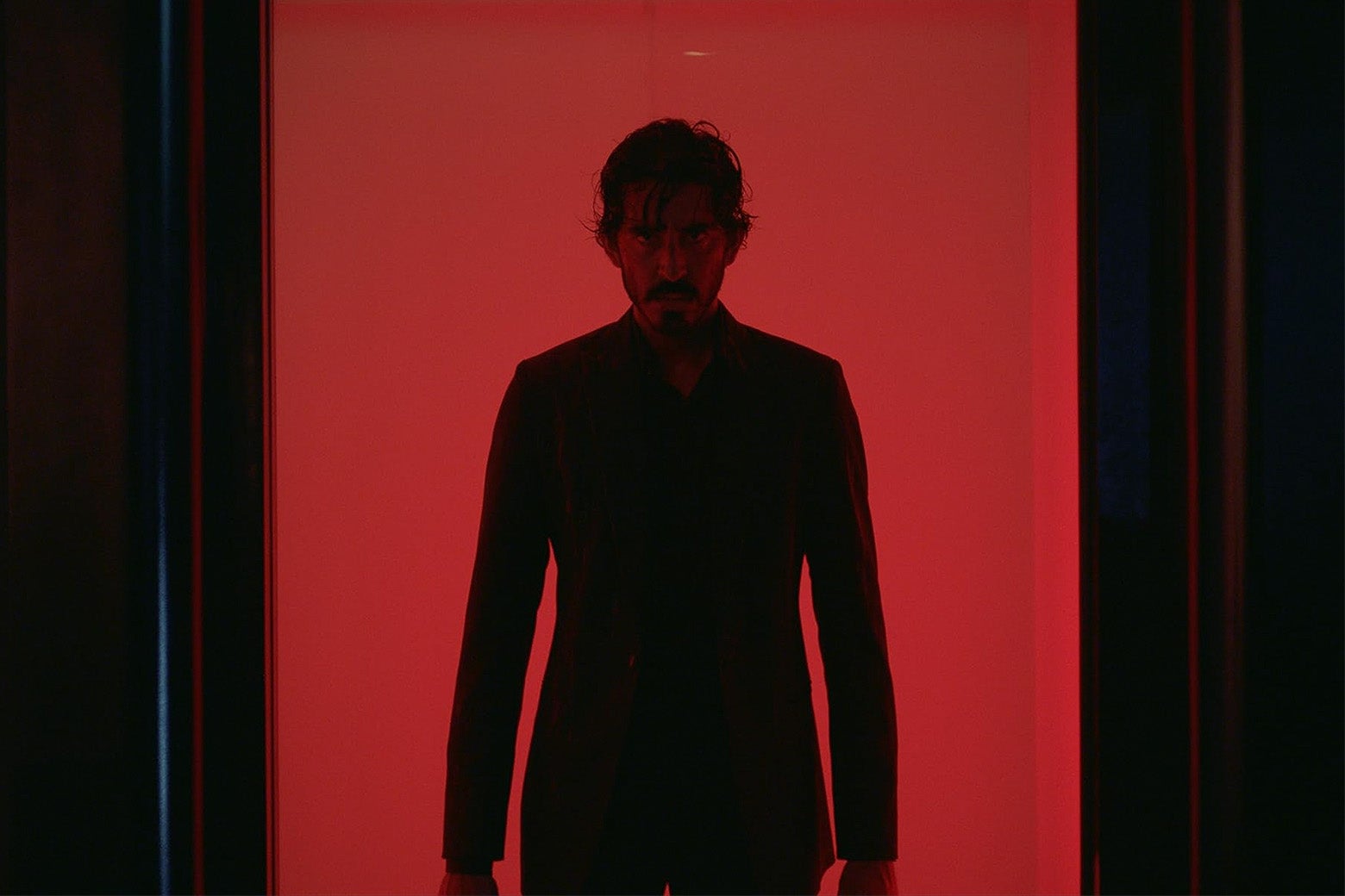 Dev Patel in a suit, standing against a red background.