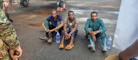 Trois jeunes hommes assis sur une chaussée, encadrés par des soldats en tenue de camouflage. Ils semblent fatigués et portent des vêtements simples, deux d’entre eux sont pieds nus et le troisième porte des sandales. Chacun tient une bouteille d’eau. Le contexte semble être après une arrestation.