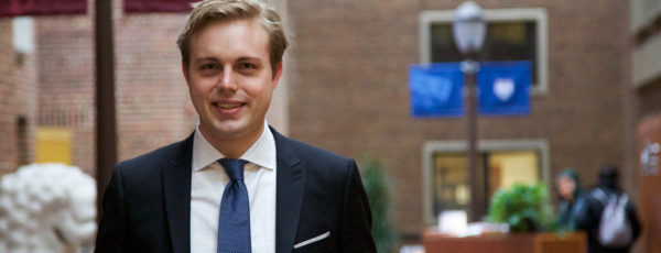 Closeup of Jon Hartley, WG'17, blonde in a navy suit, standing in the brick atrium of Steinberg-Dietrich Hall.