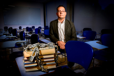 Professor Gabriel Rodriguez in a classroom standing next to stacks of books wrapped in a chain and giant padlock.