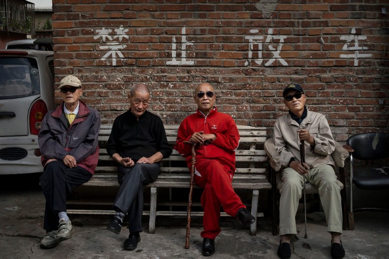 Four retired Chinese men sit on a bench outside an apartment complex for pensioners in Beijing. They wear sunglasses, hats, and comfortable clothing, and two of them hold walking canes.